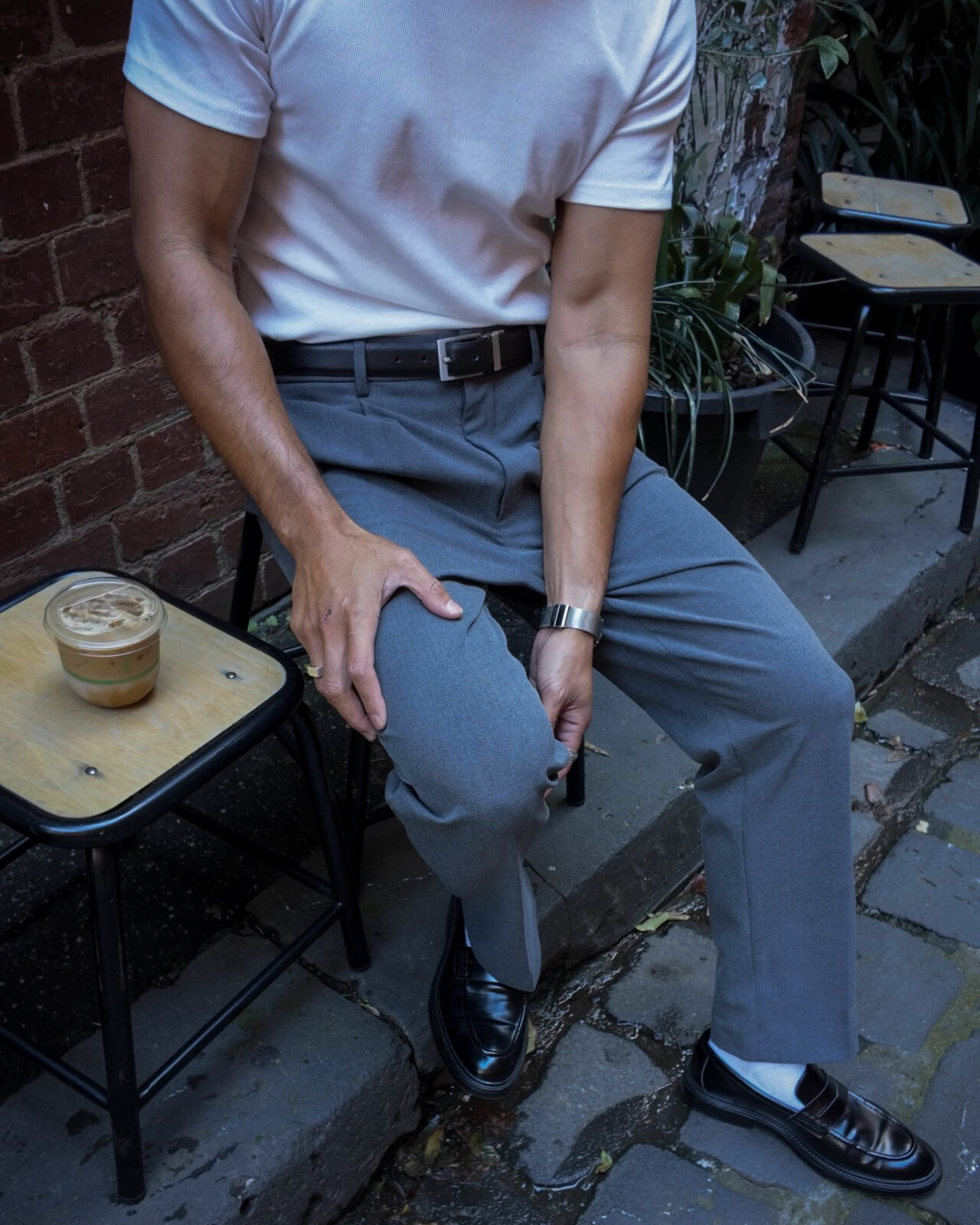 An iced coffee on a laneway stool beside grey pleated trousers and polished loafers