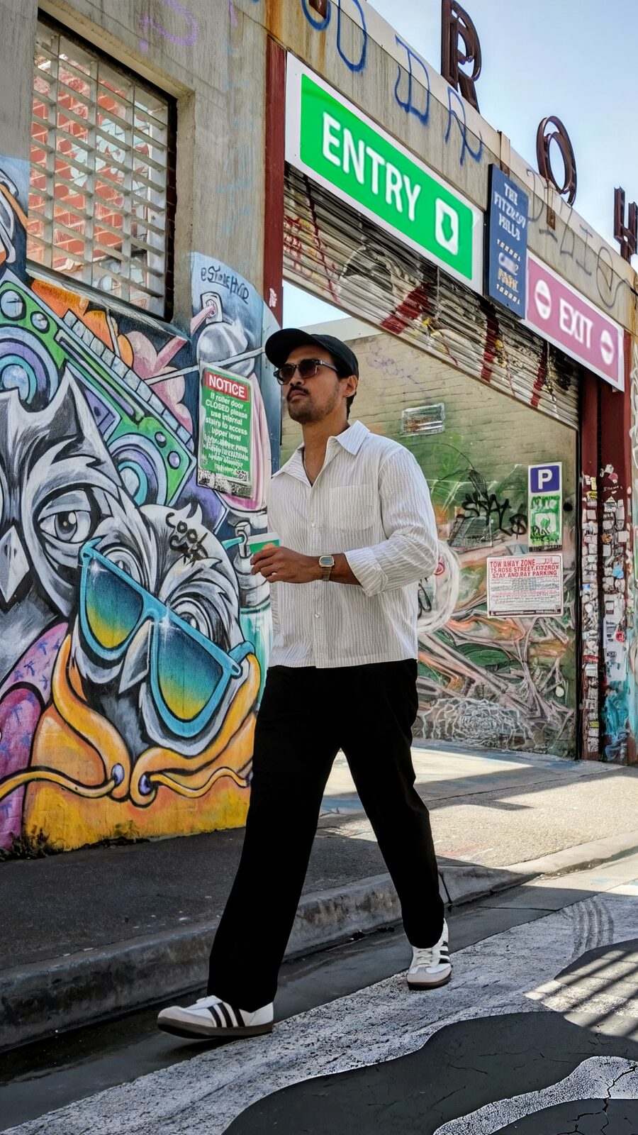 Ron walking past a graffiti-covered laneway in Fitzroy, white shirt, black trousers, coffee in hand