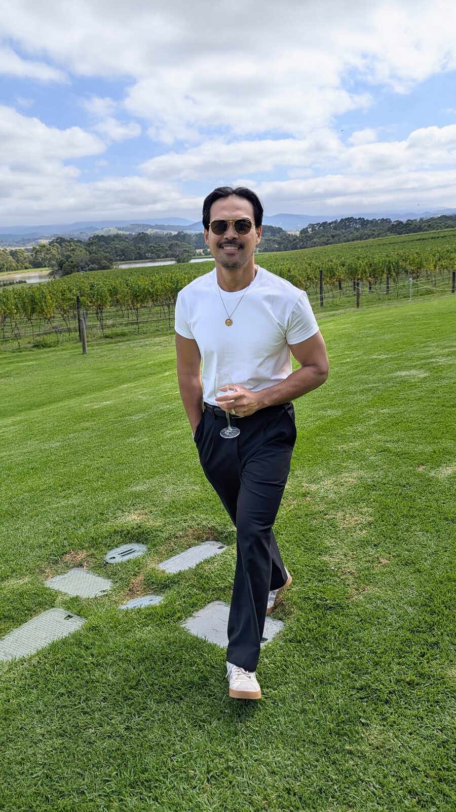 Ron walking through a Yarra Valley vineyard in a white tee and black pleated trousers, wine glass in hand